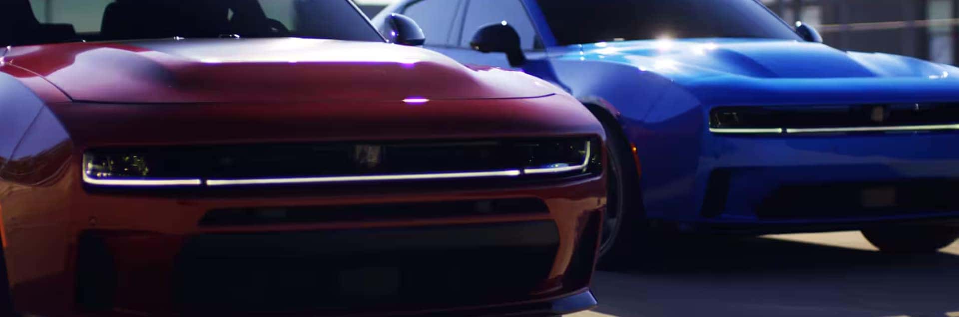 Red Dodge Charger in the foreground with a blue Charger behind it, both shown from the front with lights on.