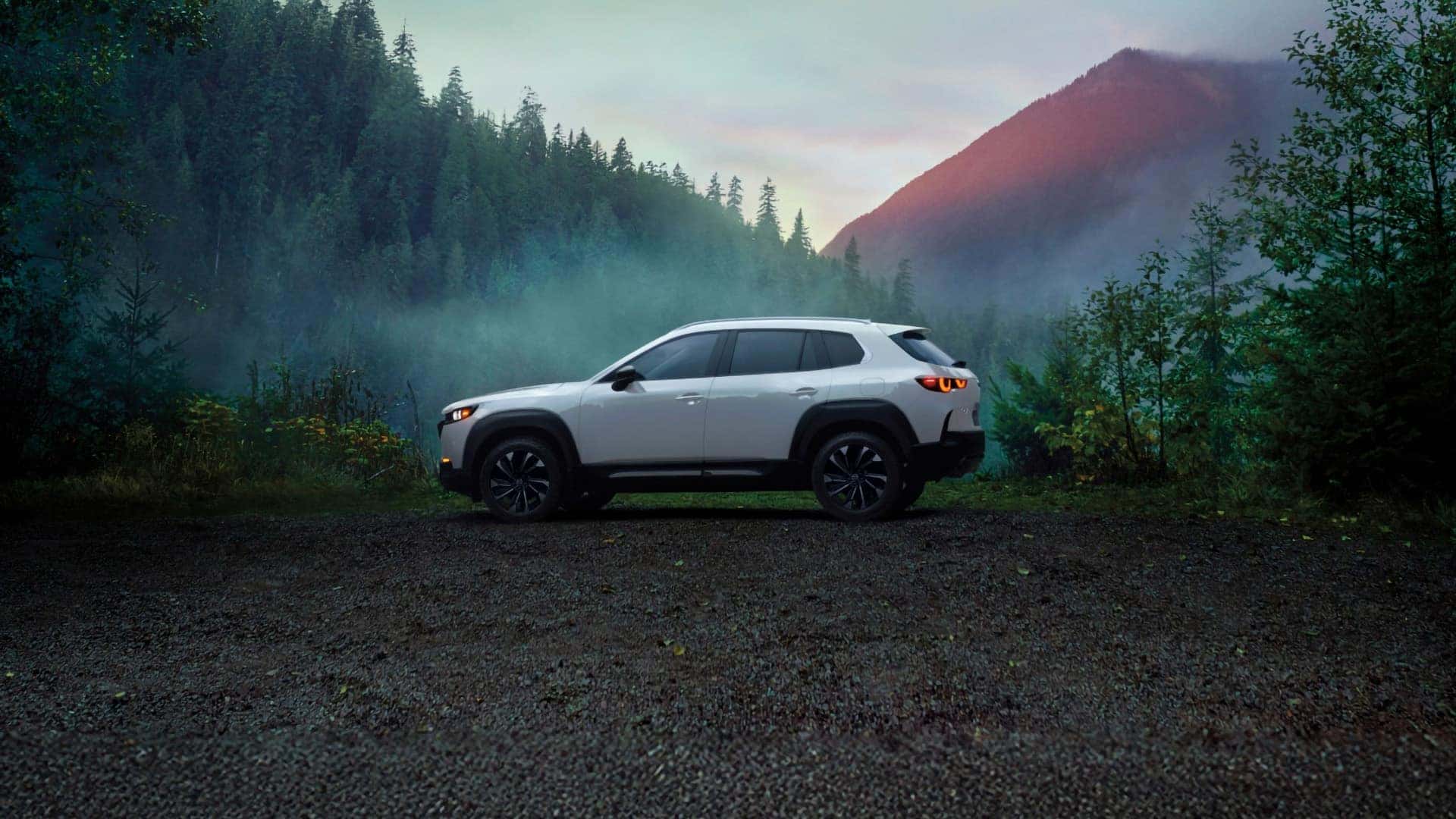 White 2026 Mazda CX-50 Hybrid in side profile with its LED exterior lights illuminated in a mountain forest