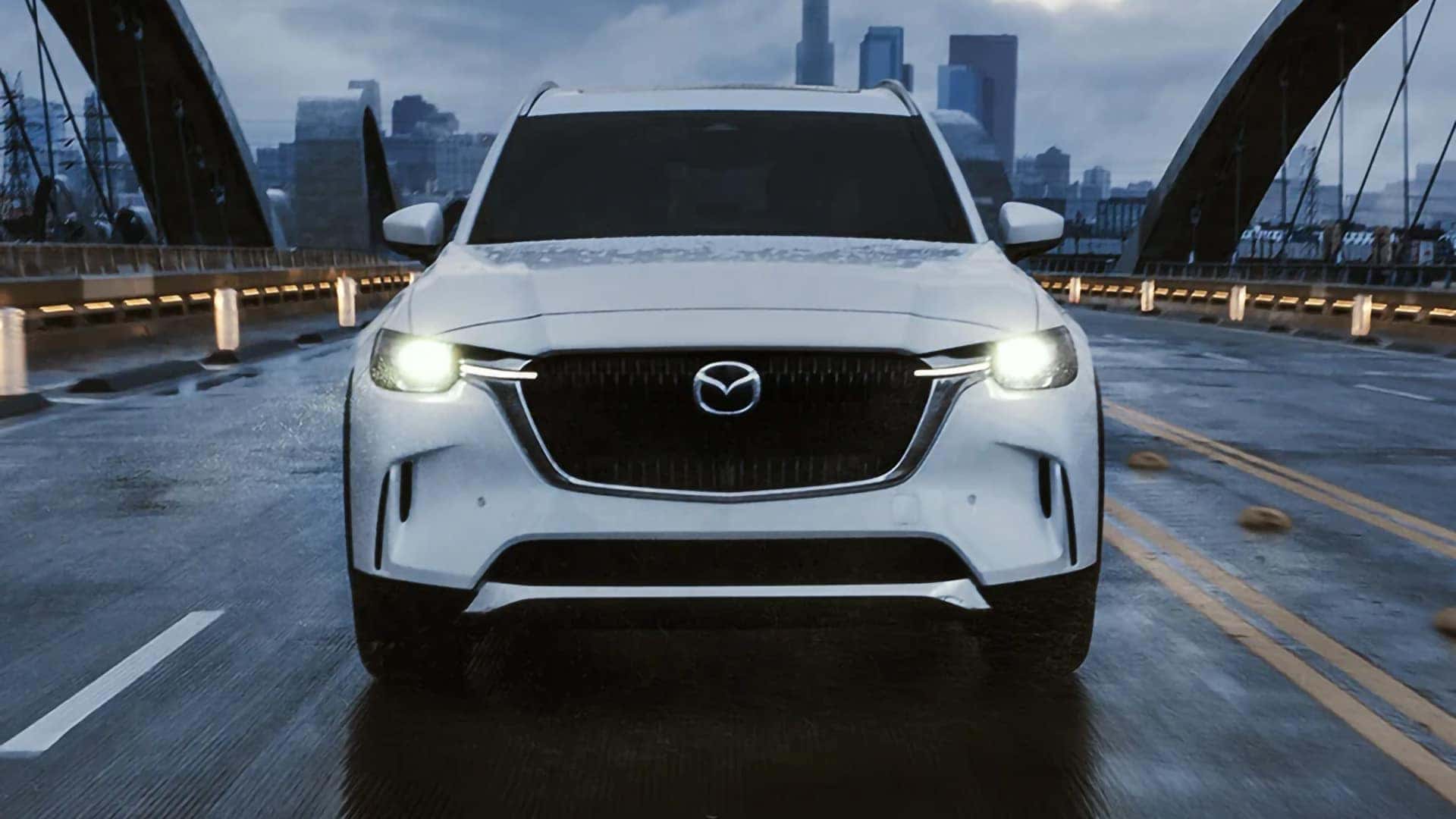 White 2026 Mazda CX-90 PHEV driving across a wet bridge with its LED headlights illuminated