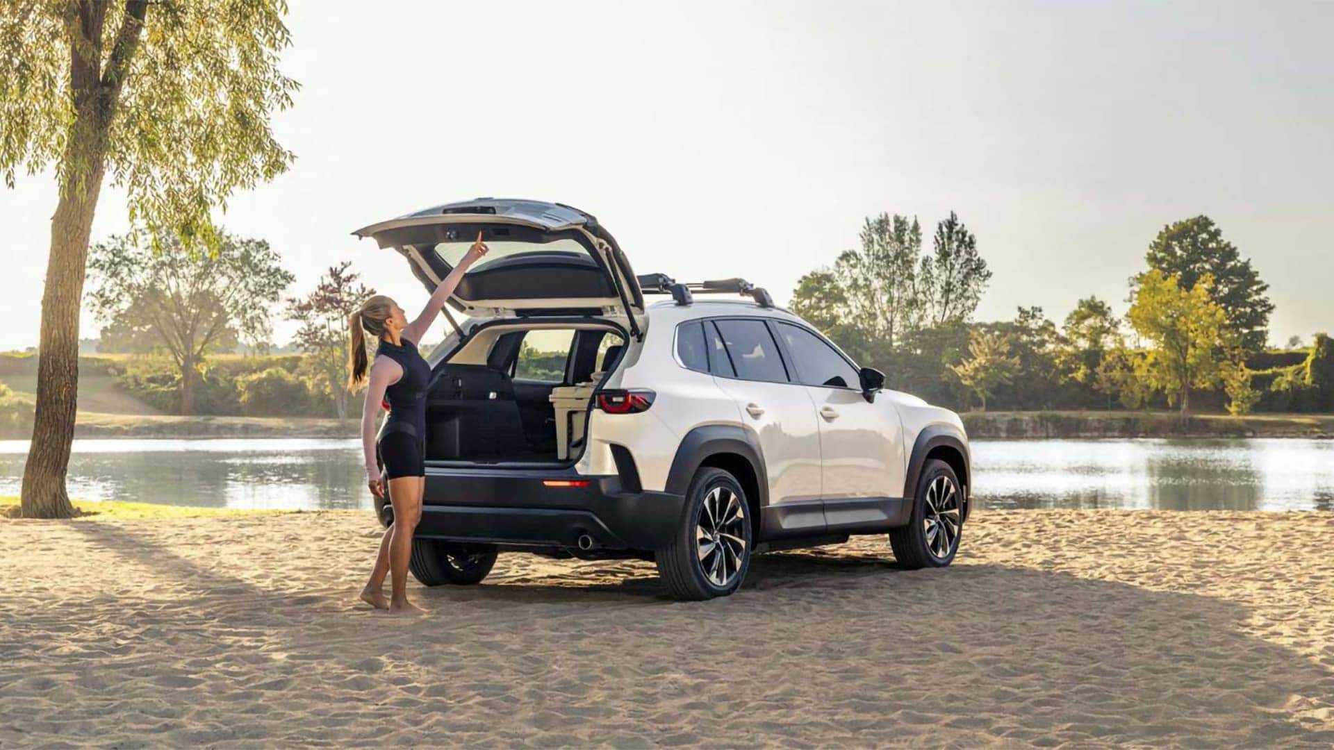 Woman opening the rear power liftgate of a white 2026 Mazda CX-50 Hybrid beside a lake
