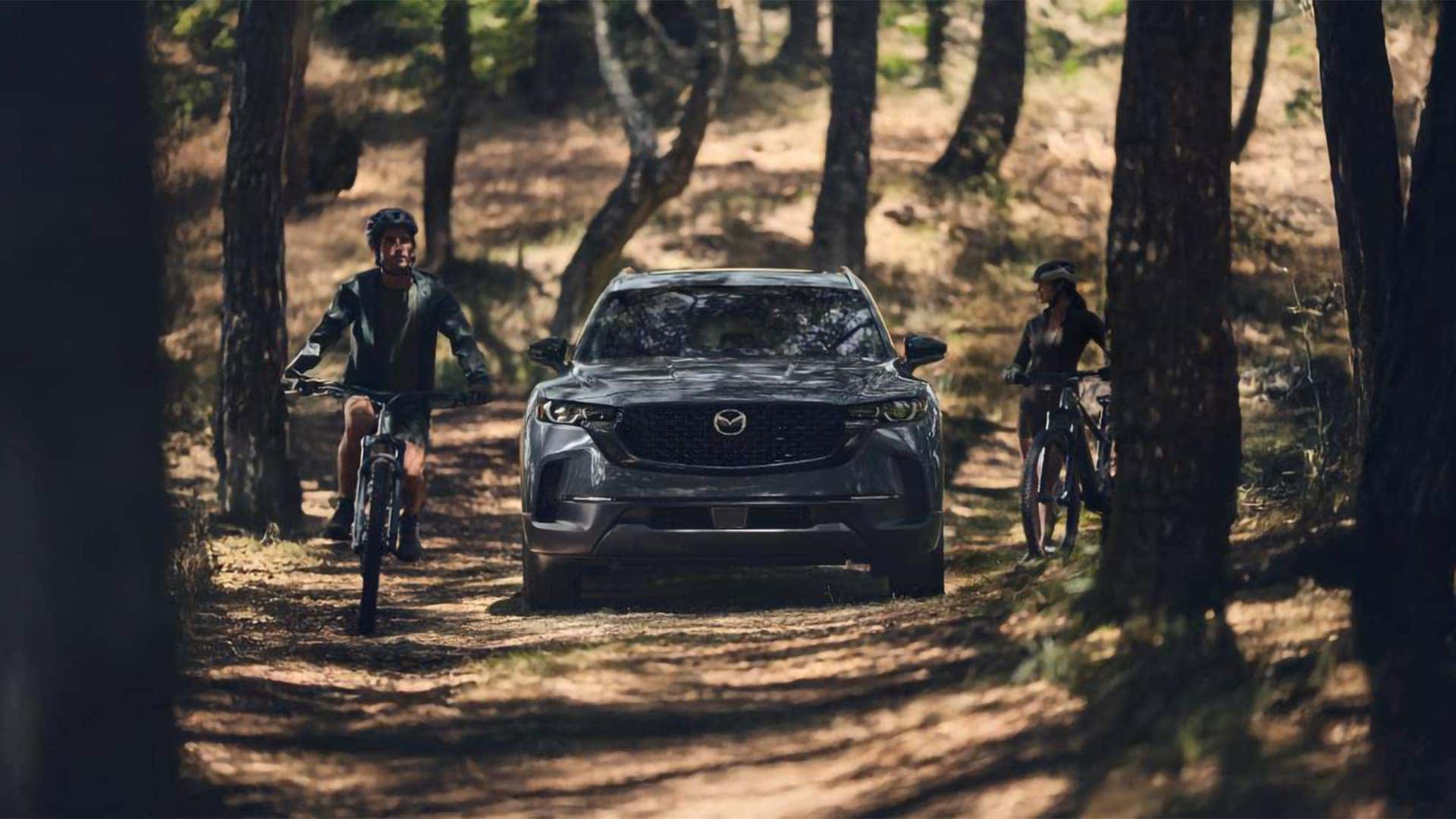 Gray 2026 Mazda CX-50 Hybrid parked on a forest trail between two cyclists