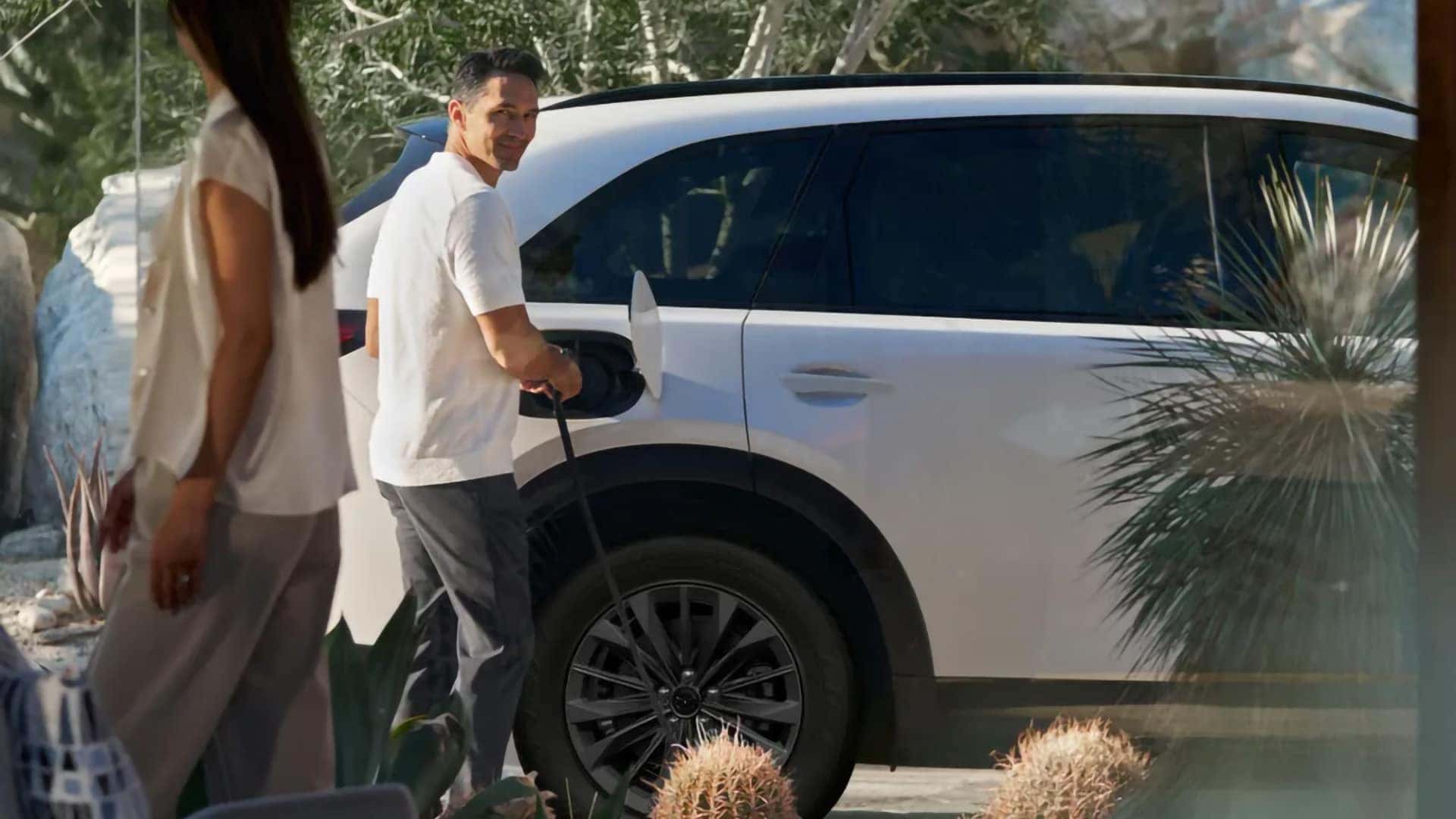 Driver connecting a charging cable to a white 2026 Mazda CX-70 PHEV outside a home