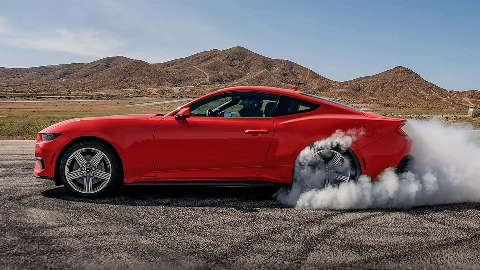 Red 2026 Ford Mustang fastback shown in profile during a burnout on an open road