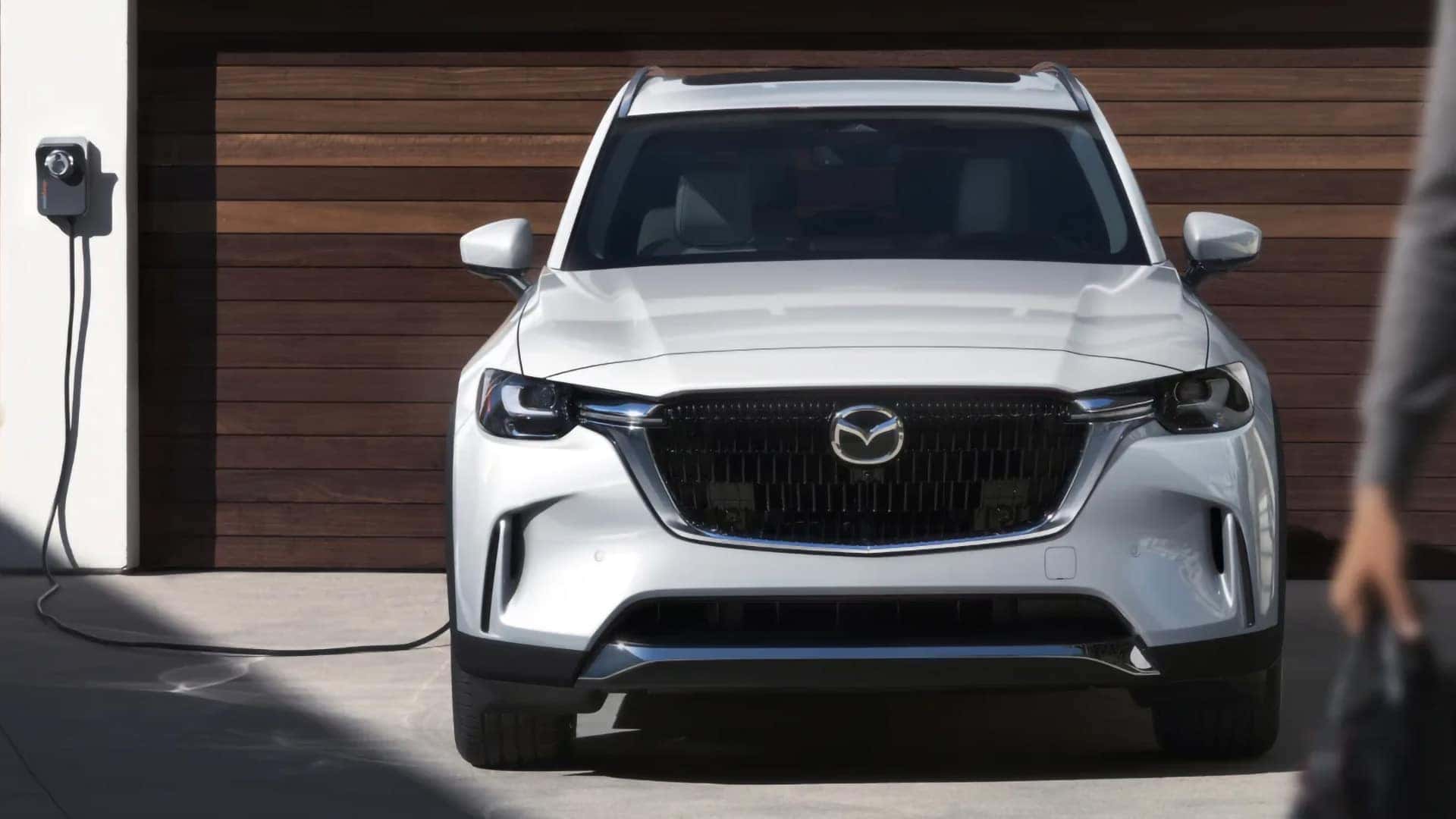 White 2026 Mazda CX-90 PHEV connected to a home charging station outside a garage