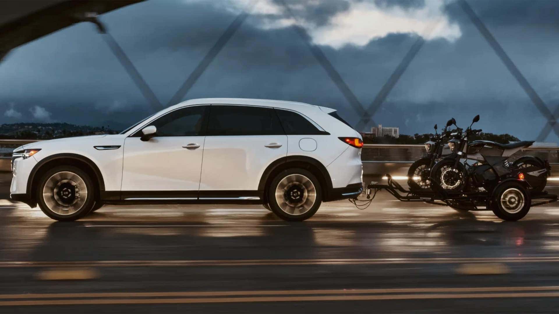 White 2026 Mazda CX-90 PHEV towing two motorcycles on a trailer across a wet bridge