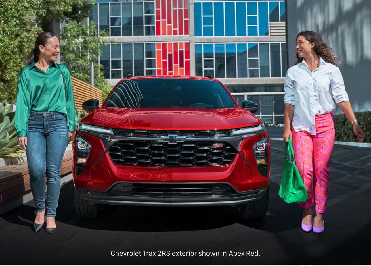 Front view of a 2026 Chevrolet Trax 2RS in Apex Red Metallic flanked by two women in colorful outfits.